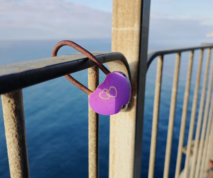 A purple heart-shaped padlock with interlocking hearts is attached to a metal railing overlooking the sea.
