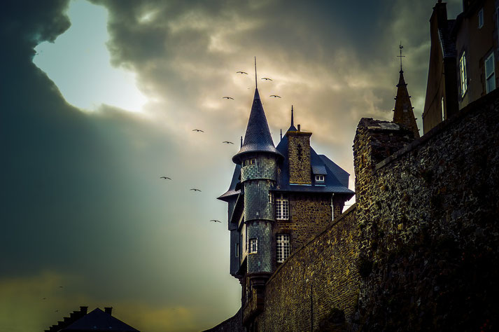 A dramatic view of a castle with pointed towers, surrounded by a stone wall. Dark, moody clouds loom overhead, and several birds are flying in the sky, adding to the eerie atmosphere.