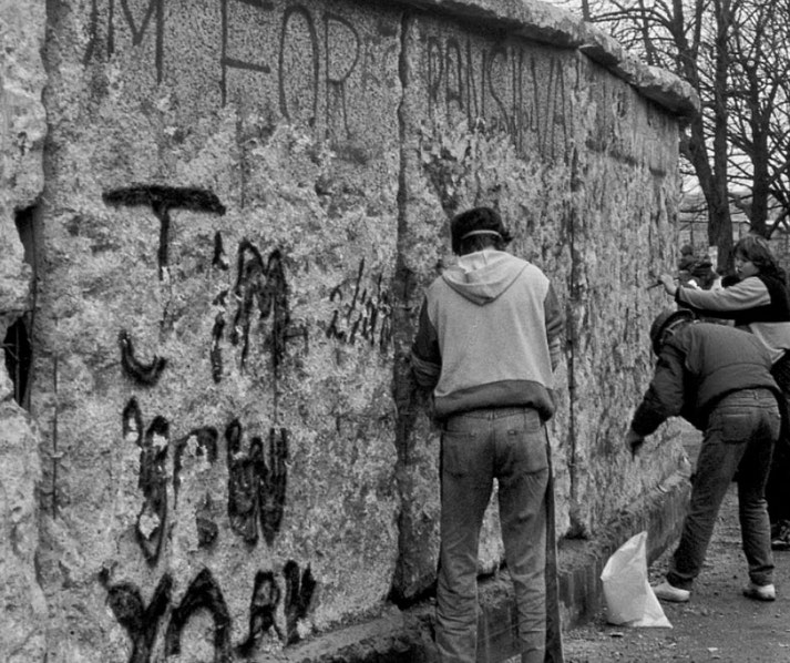 People chip away at the Berlin Wall with tools in a black and white photo capturing a moment of its fall.