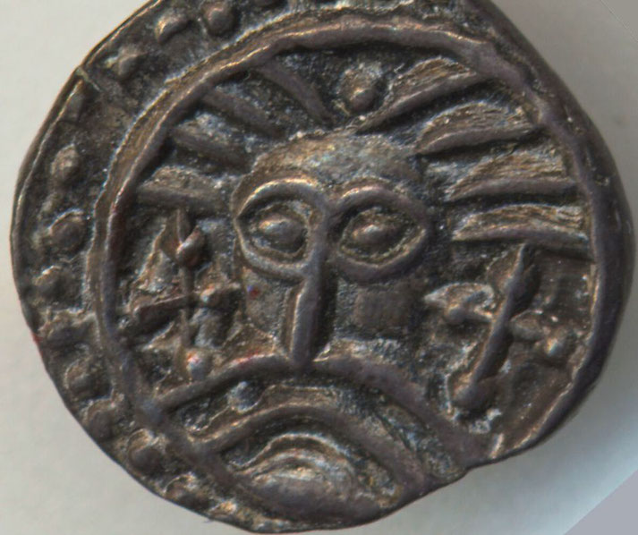 A small early medieval silver coin. The design features a stylized face with radiating lines, possibly representing a deity, ruler, or sun motif, with crosses and symbols.