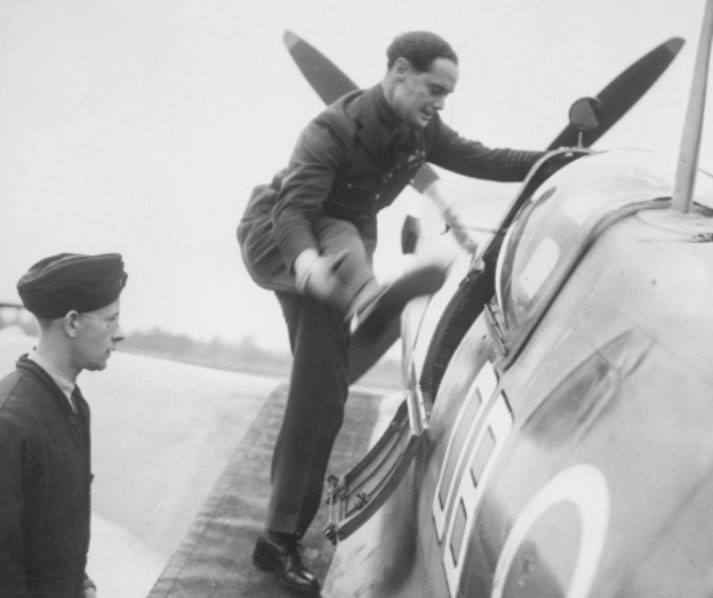 A pilot in a military uniform climbs into the cockpit of a fighter plane, possibly a Spitfire, while a ground crew member watches.
