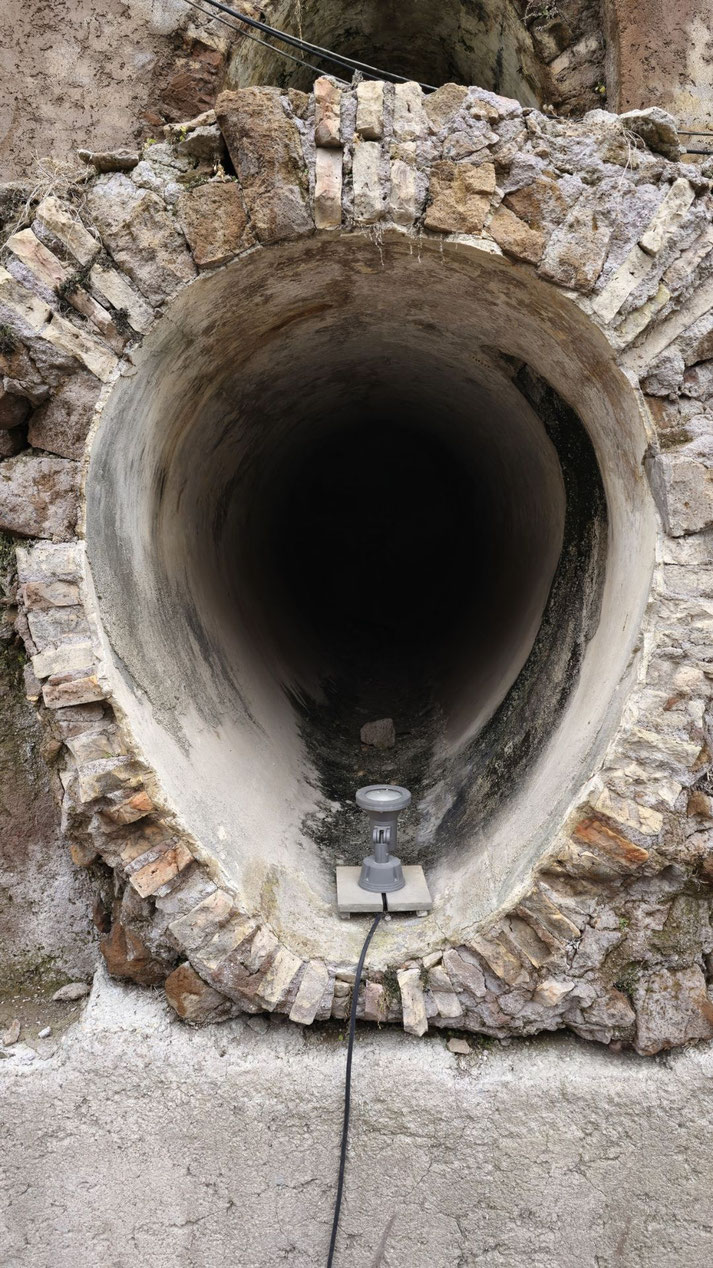 Ancient stone and brick tunnel entrance with a rounded, worn interior and a spotlight placed at the opening, likely part of a historical site or aqueduct.