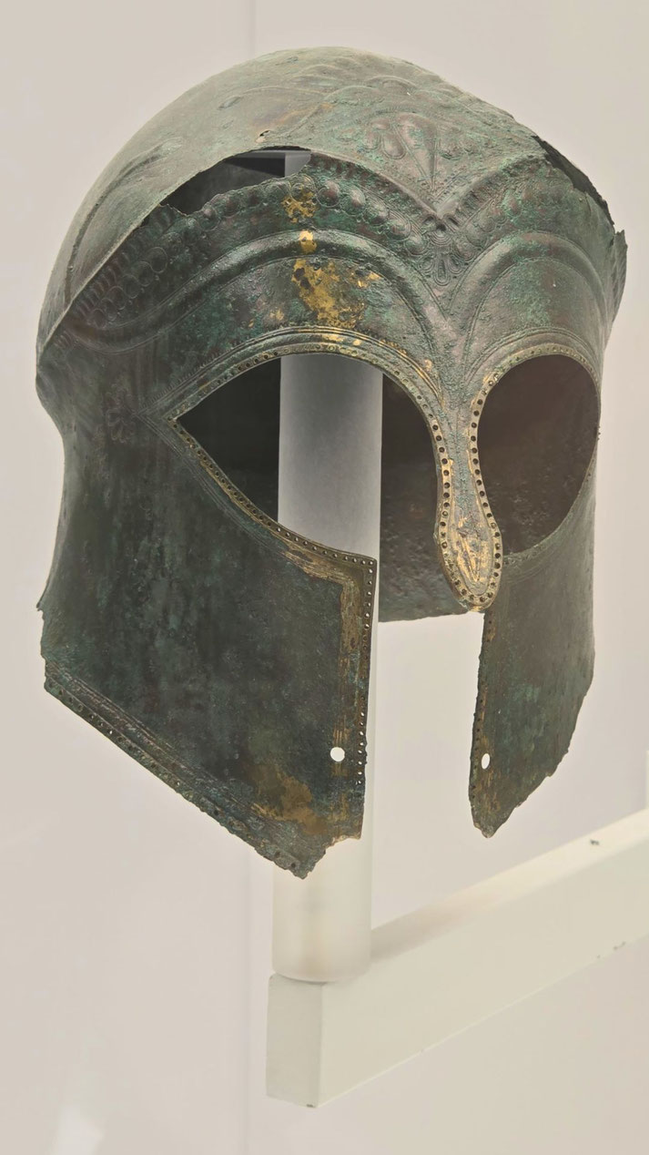 Ancient bronze helmet with ornate engravings, prominent eye openings, a curved nose guard, and edge perforations; shows signs of corrosion and age.