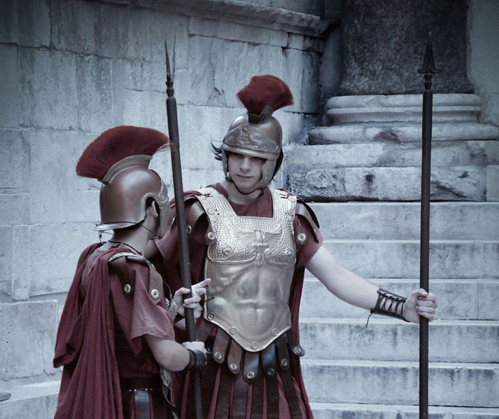 Two Roman reenactors dressed as soldiers, wearing red tunics, plumed helmets, and ornate armor. They stand guard with spears near stone steps.