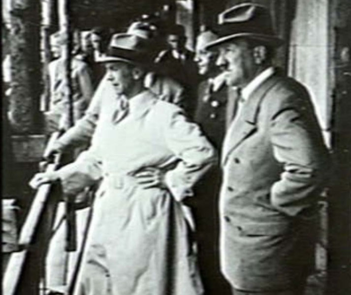 Adolf Hitler and Joseph Goebbels standing on a balcony, wearing suits and hats, observing an event below.