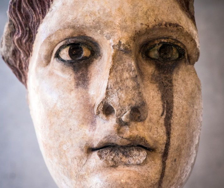 Close-up of an ancient painted statue head with wide eyes and streaked dark stains beneath them, suggesting tear-like marks.