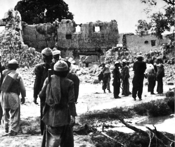 Black and white scene of armed men and civilians among rubble and collapsed stone material buildings in a devastated village after conflict.
