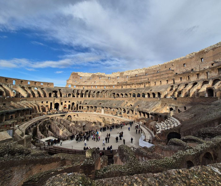 Interior view of the Roman Colosseum with tiered stone seating, exposed hypogeum, and tourists exploring the arena under a dramatic partly cloudy sky.