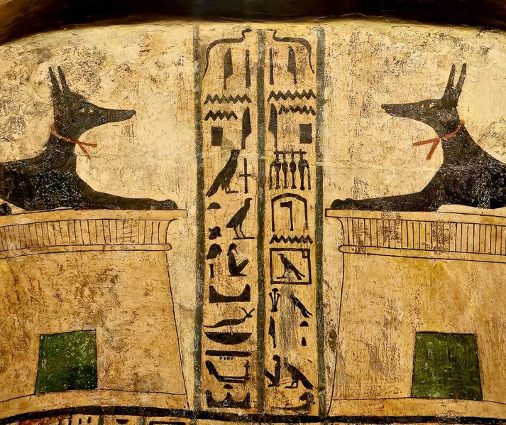An ancient Egyptian tomb painting depicting two black Anubis figures on pedestals, flanking vertical hieroglyphic inscriptions. The artwork symbolizes protection and the afterlife.