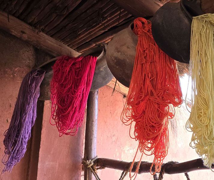 Dyed yarn in shades of purple, red, orange, and yellow hanging from rustic clay pots in a traditional setting. The scene suggests a textile dyeing or weaving process in an artisanal workshop.