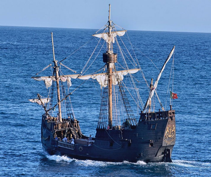 A replica of a 15th-century caravel sailing on a calm blue ocean. The ship features multiple masts, white sails, and detailed wooden craftsmanship, with a small flag at the stern.