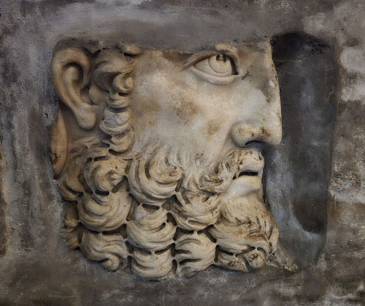 Weathered stone relief of a bearded man's profile, featuring deep-set eyes and curly hair, carved into a textured surface.