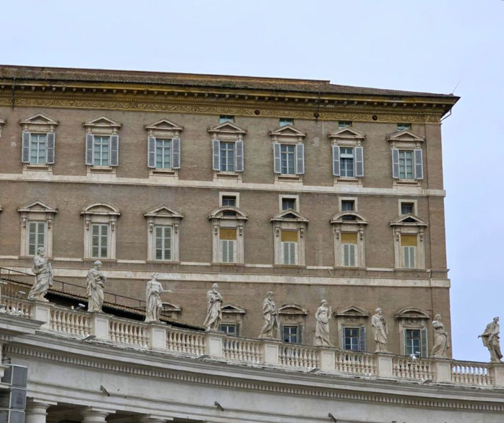 A section of the Apostolic Palace with shuttered windows and a row of statues lining the balustrade below against a cloudy sky.