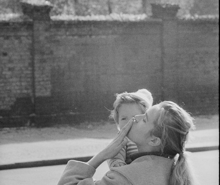 Black and white photo of a woman holding a child and blowing a kiss, standing in front of a tall brick wall.