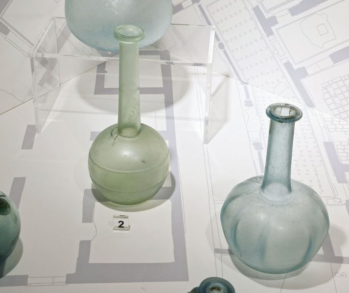 Two ancient glass bottles with long necks and bulbous bases are displayed on a museum layout map with a clear stand and numbered label.