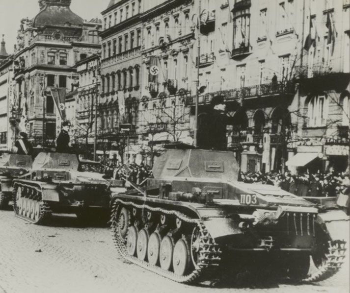 German tanks roll through a European city street during a WWII military parade, with crowds and flags lining the buildings.