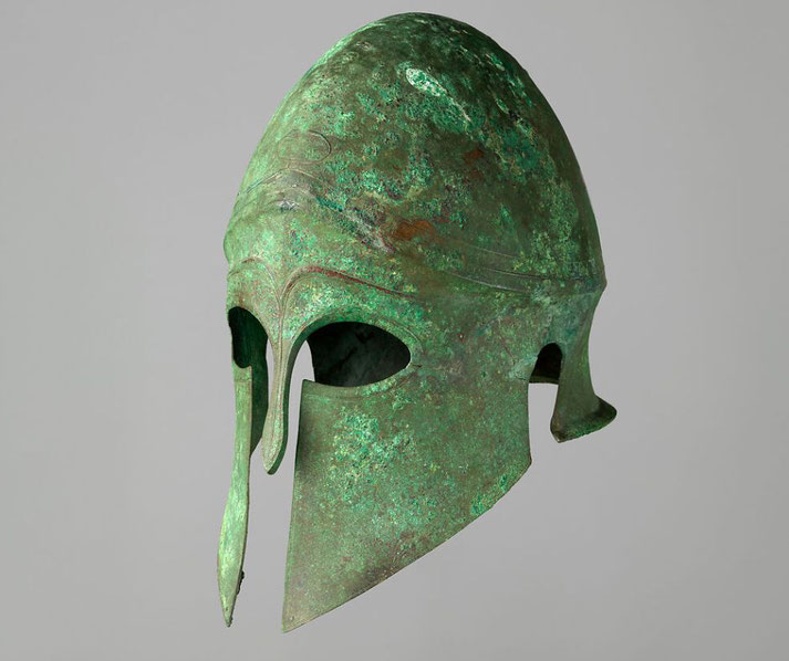 Bronze Greek helmet with elliptical eye openings and ear cutouts, paired with anatomical greaves, exemplifying late Archaic and early Classical armor.