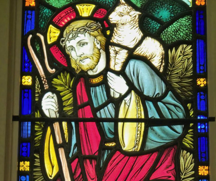 A vibrant stained glass depiction of Jesus as the Good Shepherd, carrying a lamb on his shoulders and holding a staff, surrounded by colorful patterns and religious iconography.