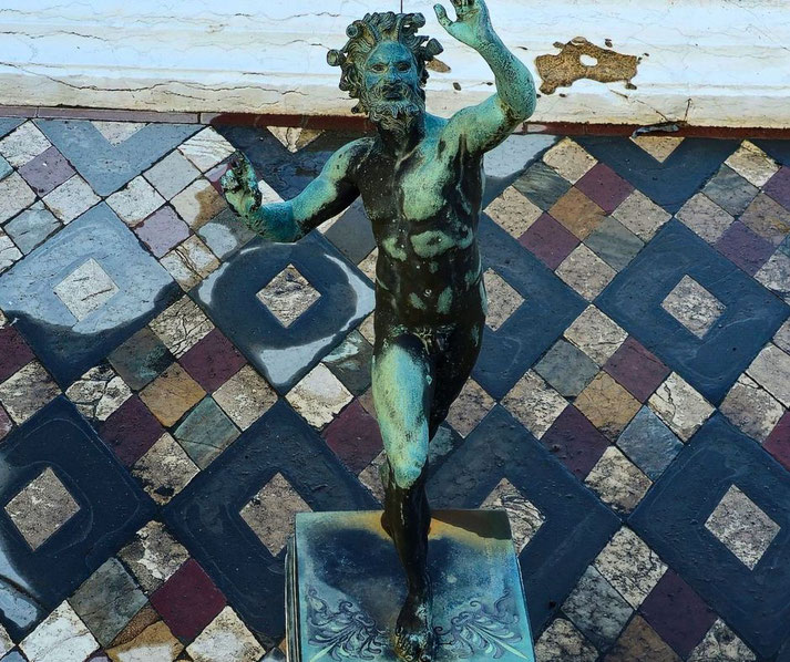 S bronze statue of a dancing faun in the House of the Faun in Pompeii, set against a colorful, geometric mosaic floor that highlights its artistic and cultural significance.