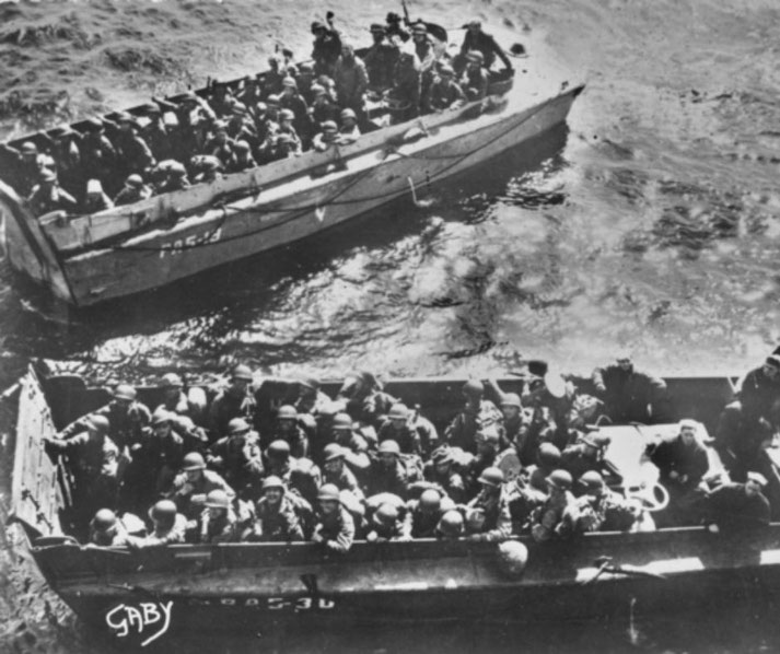 Two landing craft packed with soldiers approach shore during a WWII amphibious operation, seen from above.