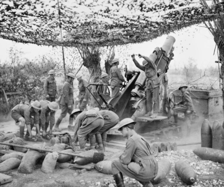 WWII soldiers operate a large artillery gun under a camouflaged net, loading shells and preparing to fire in a battlefield setting.