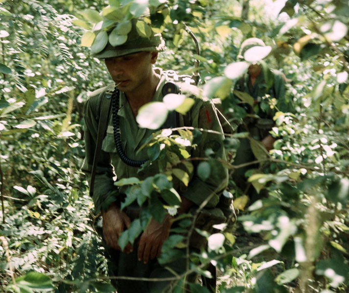 Two American soldiers move cautiously through dense jungle foliage during a patrol, partially obscured by leaves.