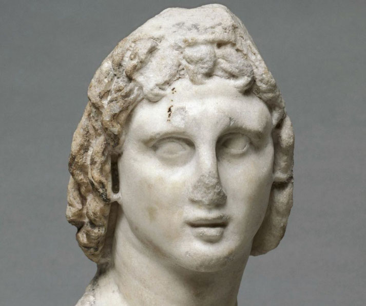 A weathered marble bust depicts a young man with curly hair and blank eyes, showing signs of age and damage, particularly on the nose and forehead.