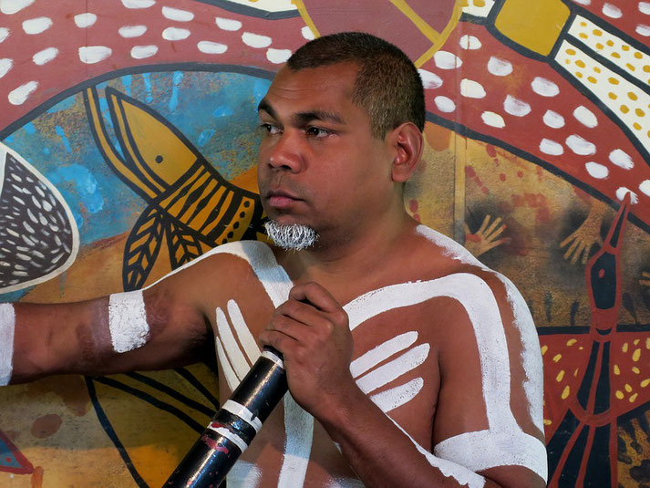 A man with traditional white body paint holds a didgeridoo in front of a colorful Aboriginal artwork backdrop.