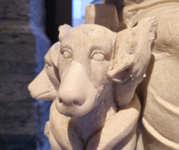 Close-up of a stone dog sculpture with a restored smooth snout, detailed ears, and simplified eyes, positioned beside a larger figure.