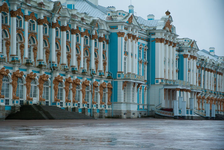 Blue and white palace with large windows, tall columns, and sculpted figures along the facade, seen on a wet, empty square.