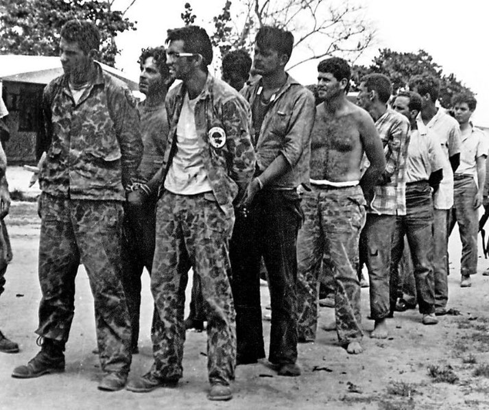 A line of captured men, some in military fatigues and others barefoot, stand with hands bound during the aftermath of a failed operation.