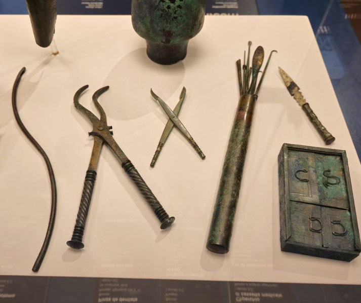 Ancient bronze medical tools including forceps, scalpel, probe set, and a case, displayed neatly on a white surface.