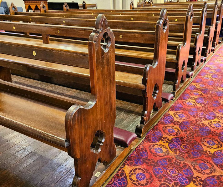 Wooden church pews with decorative cutouts are lined beside a patterned red and purple carpet aisle.