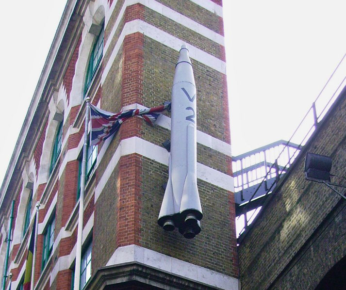 A replica V-2 rocket is mounted vertically on the corner of a brick building with a Union Jack flag beside it.
