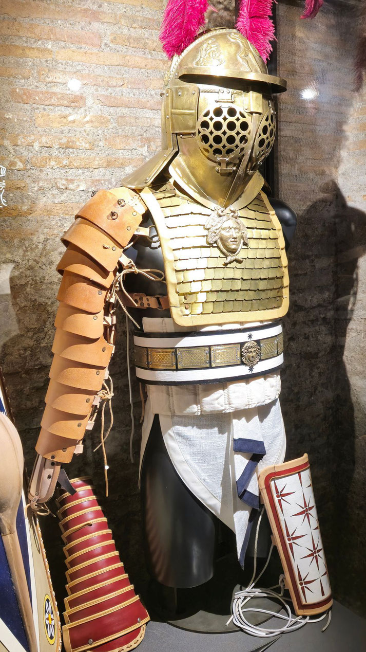 Gold helmet with perforated visor, scale chest plate bearing a Medusa emblem, leather arm guard, decorative belt, greaves and pink feather plume in a glass display case.