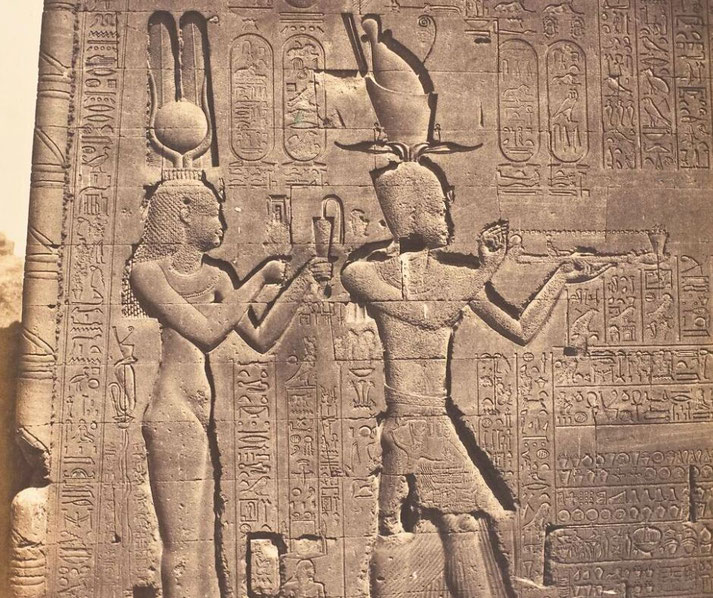 An ancient Egyptian relief depicting a pharaoh wearing a tall crown offering a tray, while a goddess, possibly Hathor, adorned with a sun disk and cow horns, holds a scepter. 