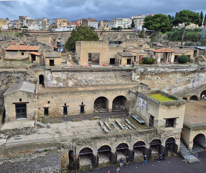 Ancient stone ruins of Herculaneum spread across terraced levels beneath a cloudy sky, with a row of arched boatsheds at the base once used to shelter vessels.