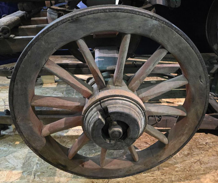 A close-up of a wooden wagon wheel with a central hub, sturdy spokes, and signs of wear, likely from an old cart or carriage used in historical transport.