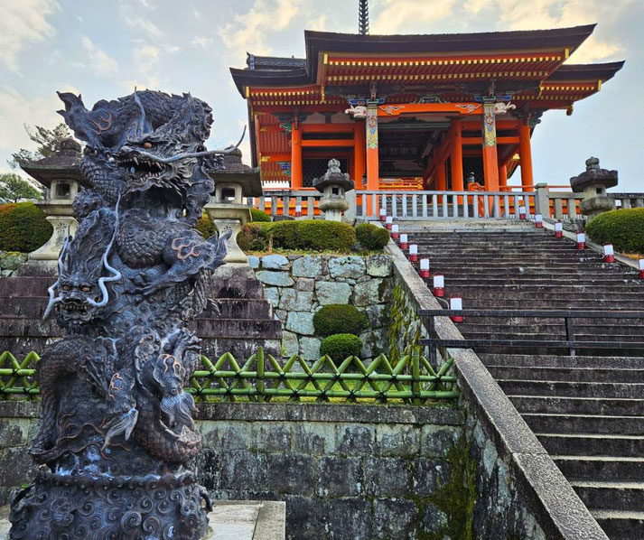 A striking dragon statue with intricate details stands in front of a traditional Japanese temple with vibrant orange pillars, ornate carvings, and stone lanterns.
