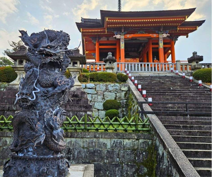 A striking dragon statue with intricate details stands in front of a traditional Japanese temple with vibrant orange pillars, ornate carvings, and stone lanterns.