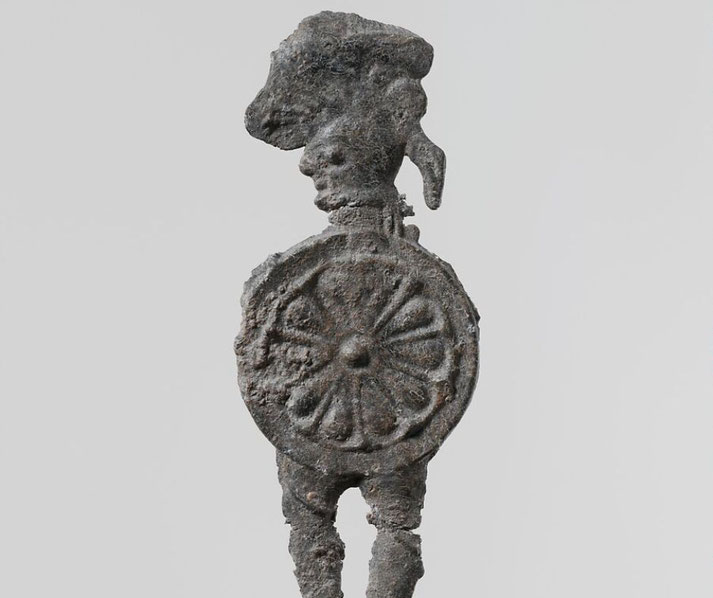 An ancient lead figurine of a warrior wearing a helmet and carrying a round shield with a radial design.