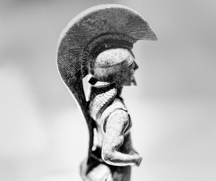 Close-up of an ancient warrior figurine with a tall crested helmet, braided hair, and muscular arms, shown in profile against a blurred background.
