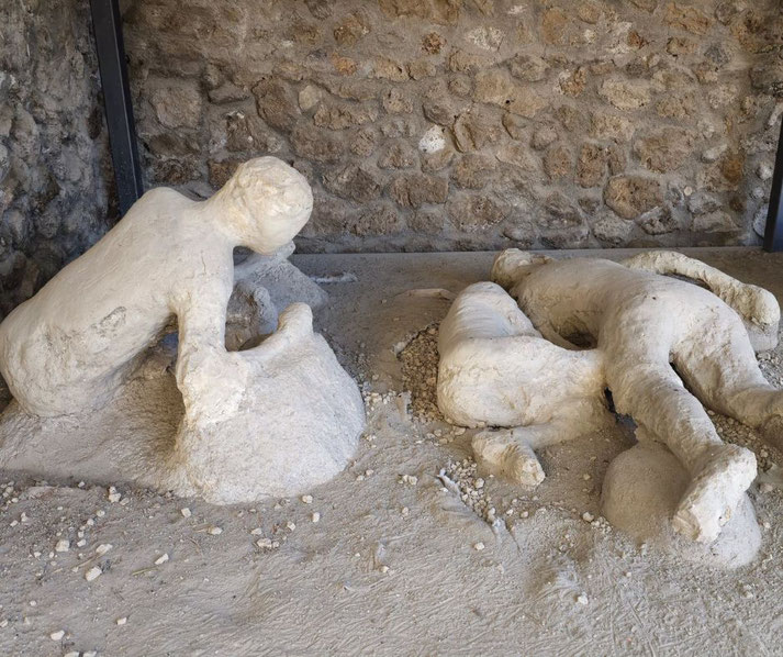 Plaster casts of victims from Pompeii's Garden of the Fugitives, capturing the tragic final moments of individuals during the eruption of Mount Vesuvius, set against a stone wall background.