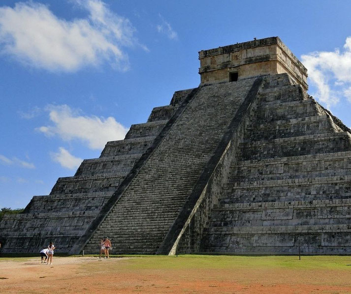 The stepped pyramid of Chichén Itzá stands tall under a bright sky, with tourists gathered at its base.