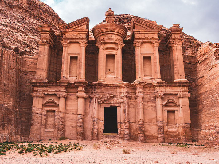 The Monastery (Ad Deir) in Petra, Jordan, an impressive Nabatean structure carved into rose-red sandstone cliffs, featuring ornate columns, a central dome, and intricate details under a cloudy sky.
