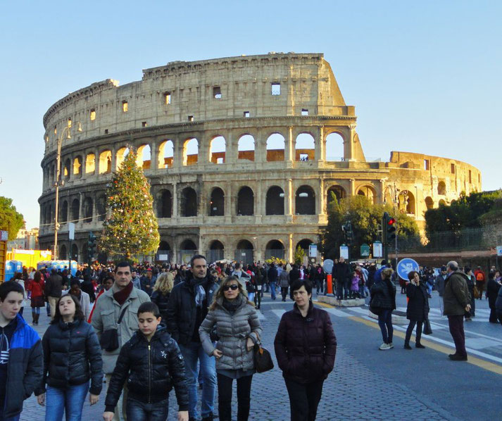 Colosseum in Rome, Italy, bustling with visitors. A decorated Christmas tree stands in the foreground, adding a festive touch to the iconic ancient amphitheater under a clear sky.
