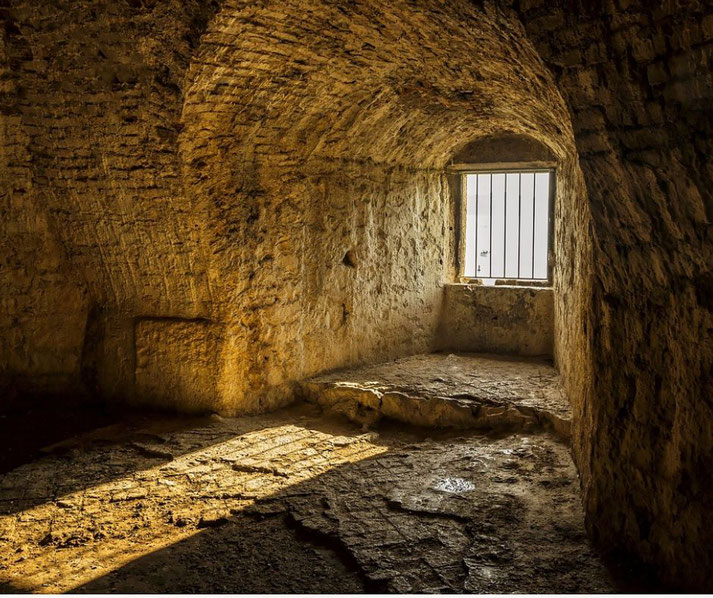 A dimly lit, ancient stone cell with an arched ceiling and a small barred window, allowing sunlight to cast shadows on the rough, uneven floor.