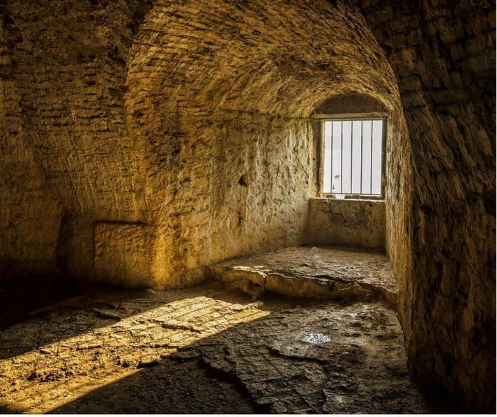 A dimly lit, ancient stone cell with an arched ceiling and a small barred window, allowing sunlight to cast shadows on the rough, uneven floor.