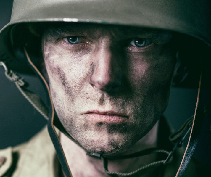 Close-up of a soldier's face with a serious expression, dirt-covered skin, and a combat helmet, capturing intensity and focus.