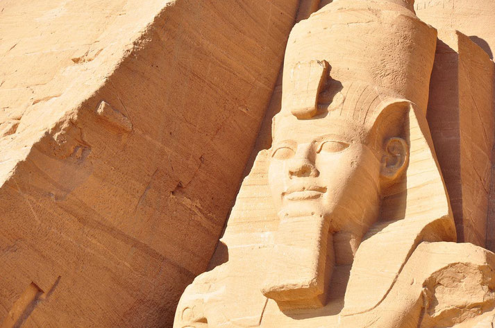 Close-up of a colossal sandstone statue of an ancient Egyptian pharaoh wearing a headdress, carved into a rock face.